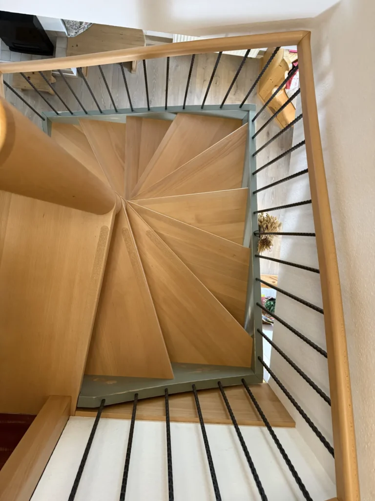 Draufsicht auf eine moderne Holzspindeltreppe mit schwarzem Metallgeländer und hellen Holzstufen, die ein geometrisches, fächerartiges Muster bilden. Die Treppe führt in einer engen Spirale innerhalb eines Holzrahmens nach unten.
