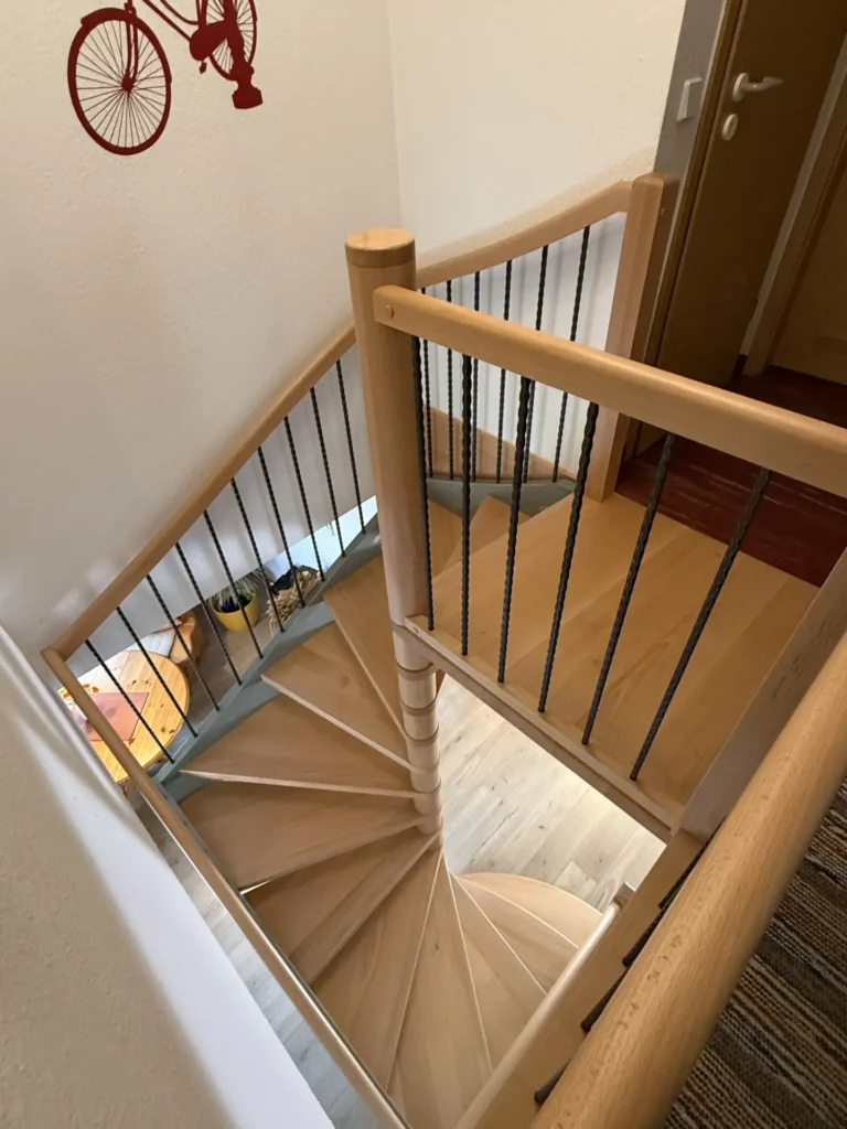 Draufsicht auf eine hölzerne Wendeltreppe mit schwarzem Metallgeländer, die in eine untere Etage führt. An der weißen Wand neben der Treppe ist ein rotes Fahrrad zu sehen.