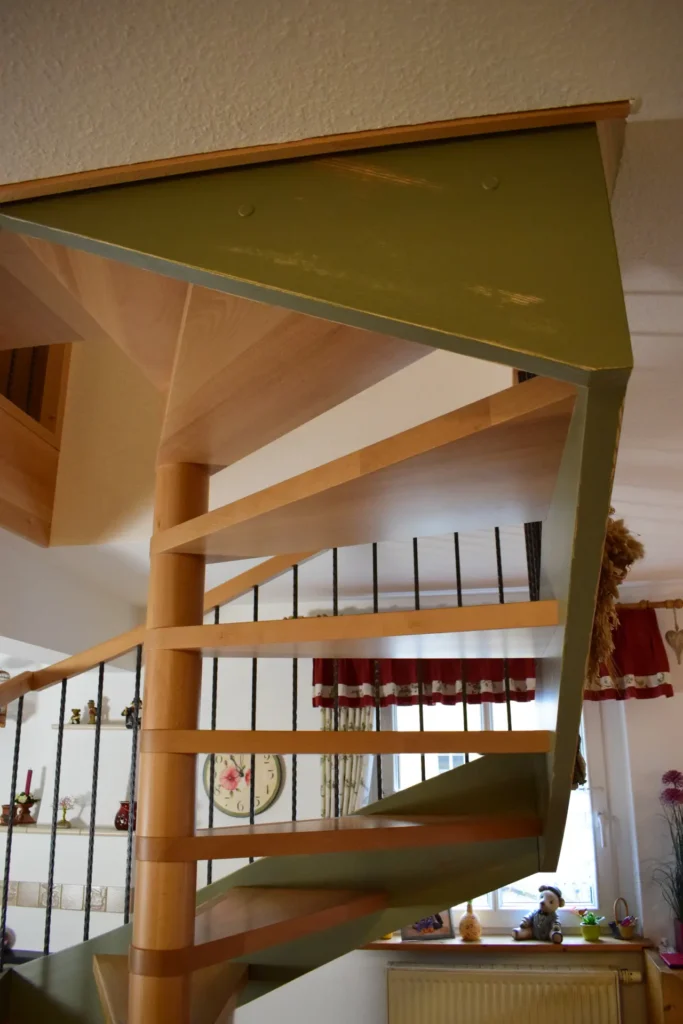 Eine spiralförmige Holztreppe mit grünen Seitenwänden führt nach oben in einen gemütlichen Raum mit Regalen, einer Wanduhr und einem Fenster mit roten Vorhängen im Hintergrund.