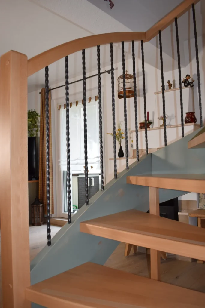 Eine Holztreppe mit hellbraunen Stufen und schwarzen Metallspindeln schlängelt sich in einem gemütlichen, gut beleuchteten Raum mit Pflanzen, Regalen und Dekorationsgegenständen im Hintergrund nach oben.