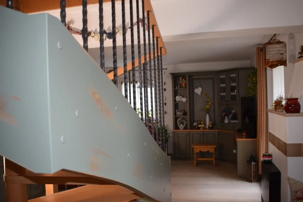 Ein gemütliches Interieur mit einer Holztreppe mit Eisengeländer im Vordergrund und einem mit Schränken, Regalen, Pflanzen und verschiedenen Einrichtungsgegenständen dekorierten Wohnbereich im Hintergrund.