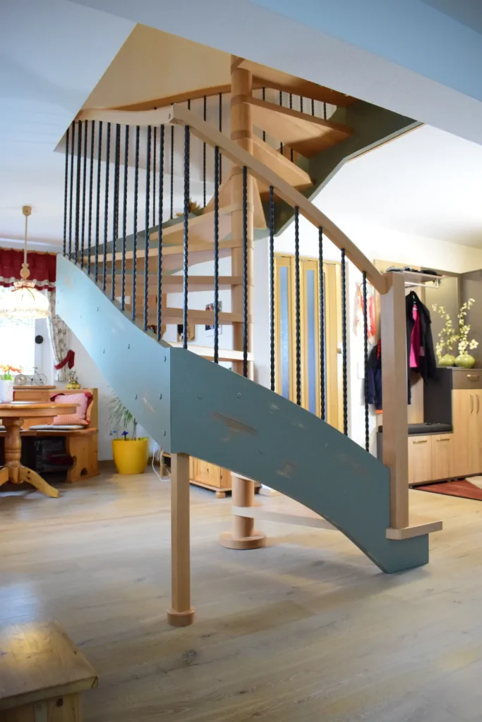 Eine moderne Spindeltreppe mit Holzstufen und blauen Seitenwänden mit schwarzem, vertikalem Metallgeländer in einem hellen, gemütlichen Wohnbereich mit Holzmöbeln und Dekorelementen.