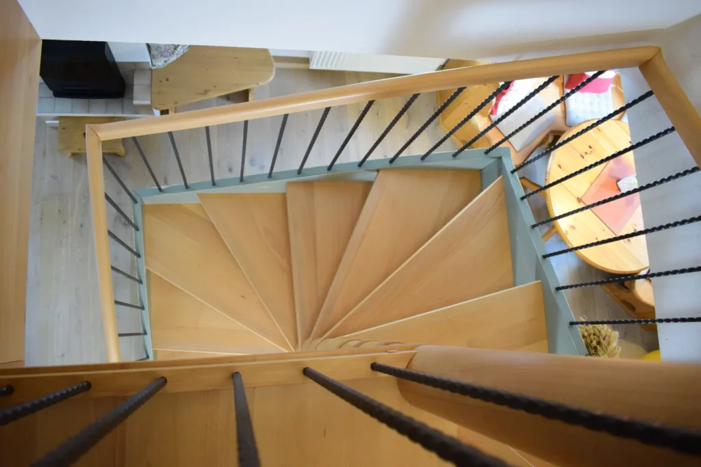 Blick von oben auf eine hölzerne Wendeltreppe mit schwarzem Metallgeländer, die in einen hellen Raum mit Holzmöbeln und einer gemütlichen Atmosphäre führt.