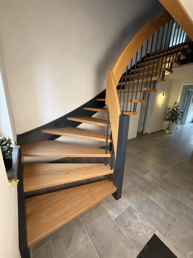 Eine moderne Wendeltreppe mit hellen Holzstufen und schwarzem Metallgeländer führt in einem hellen, gefliesten Flur hinauf, in dessen Hintergrund eine Topfpflanze und eine Wandlampe stehen.