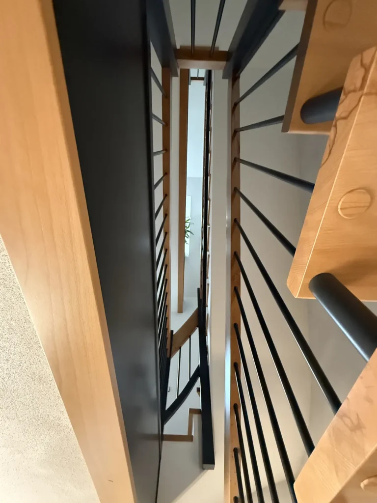Blick auf eine moderne, mehrstöckige Treppe mit Holzstufen, schwarzem Geländer und einer Pflanze, die oben durch die zentrale Öffnung sichtbar ist.