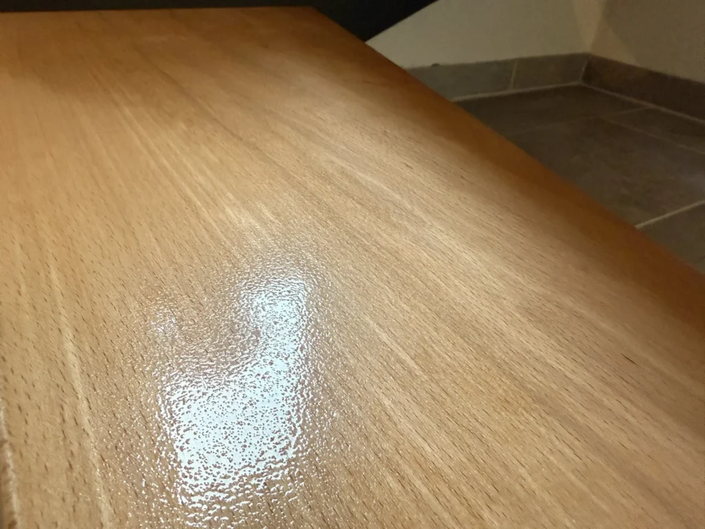 Nahaufnahme einer Holzoberfläche mit glänzender Oberfläche, die das Licht reflektiert. Die Holzmaserung ist sichtbar, und der Hintergrund zeigt einen Teil eines gefliesten Bodens und einer Wand.