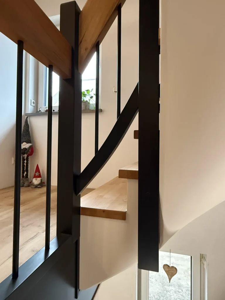 Eine Nahaufnahme einer modernen Holztreppe mit schwarzem Metallgeländer. Im Hintergrund stehen zwei Zierzwerge auf dem Boden und ein herzförmiges Ornament hängt an einem Fenster. Eine Topfpflanze steht auf der Fensterbank.