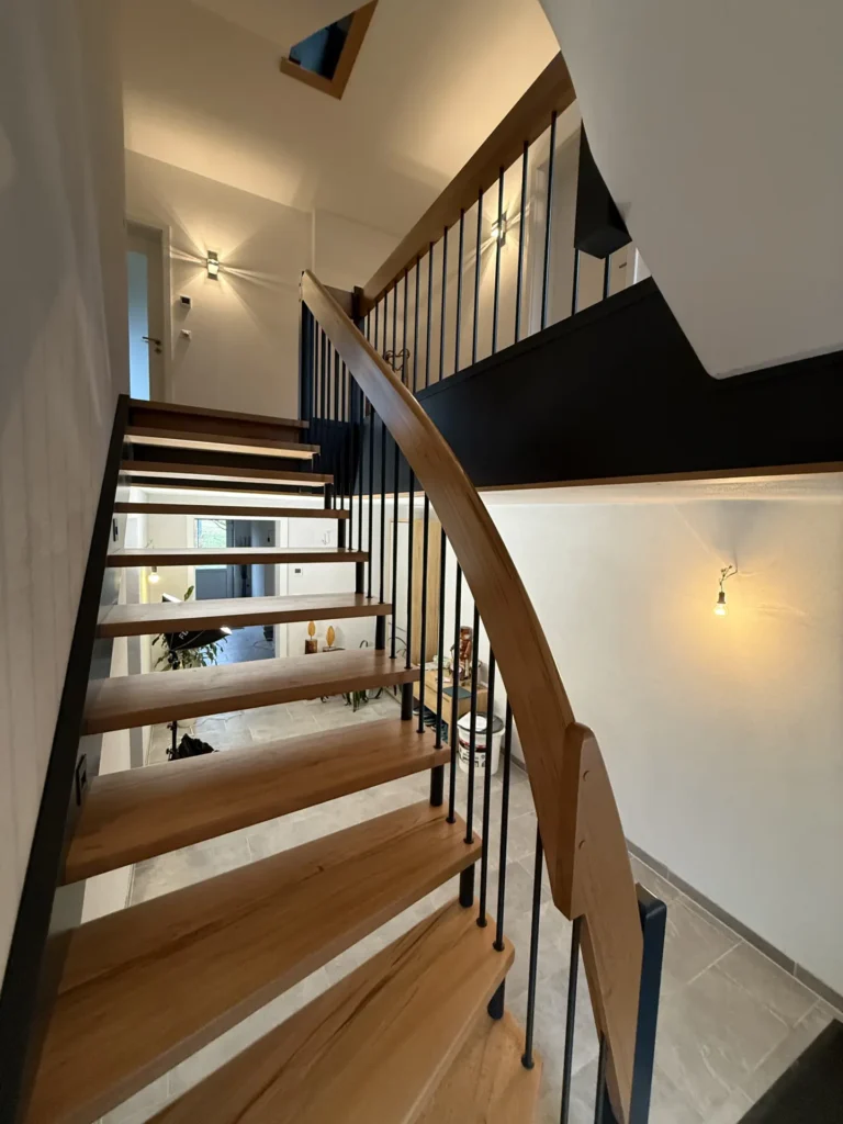 Moderne Holztreppe mit schwarzem Metallgeländer in einem gut beleuchteten Wohnbereich. Die Treppe führt von einem gefliesten Boden nach oben, im unteren Flur sind Wandleuchten und eine kleine Pflanze zu sehen.