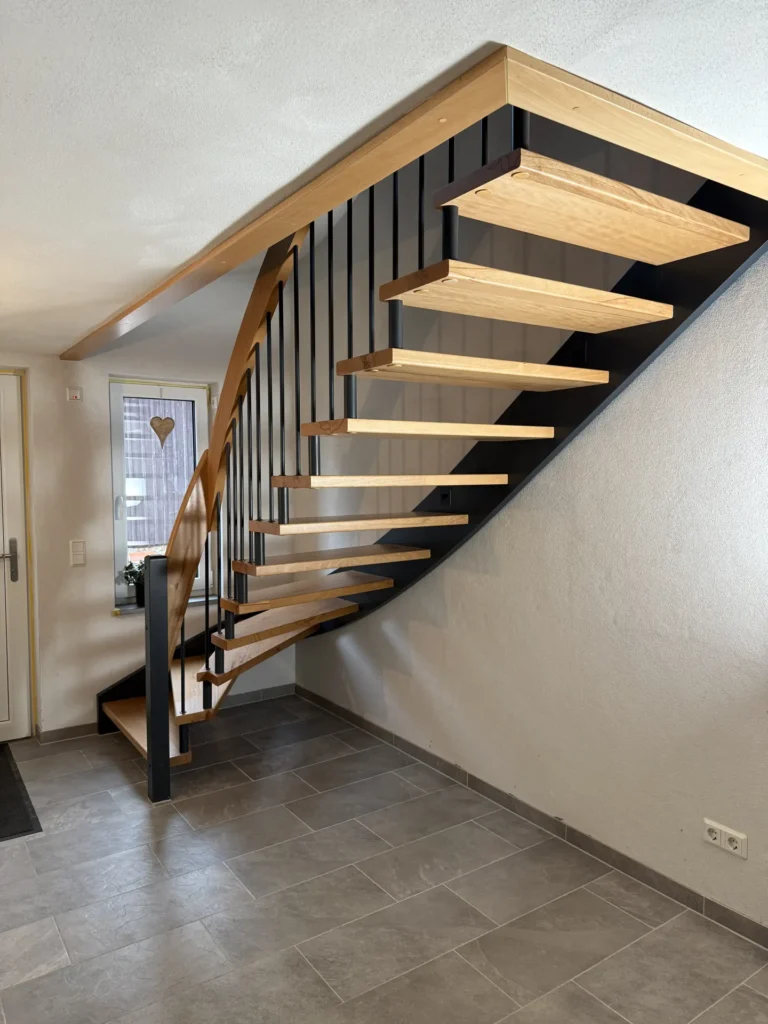 Eine moderne Holztreppe mit offenen Setzstufen und schwarzen Metallstützen führt an einer weißen Wand entlang in einen Raum mit grauem Fliesenboden und einem mit einem Herz-Ornament verzierten Fenster.