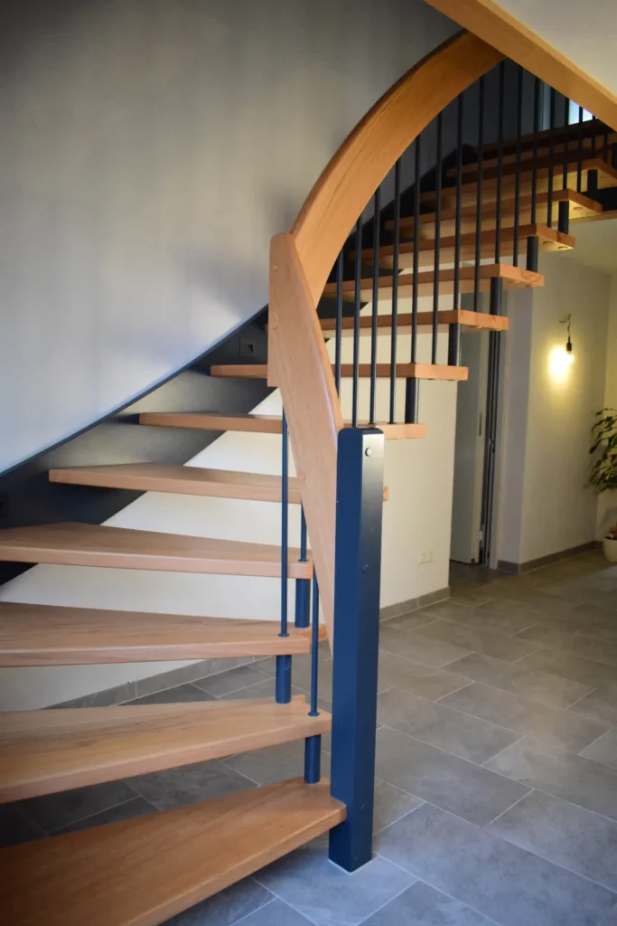 Eine moderne Holzspindeltreppe mit schwarzem Metallgeländer vor einer hellgrauen Wand und einem grauen Fliesenboden in einem zeitgenössischen Wohnbereich.