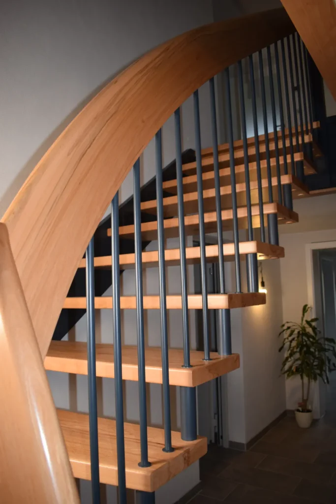 Eine moderne Wendeltreppe mit hellen Holzstufen und schwarzem vertikalen Metallgeländer schlängelt sich in einem hellen Innenraum nach oben; am unteren Ende der Treppe steht eine Topfpflanze.
