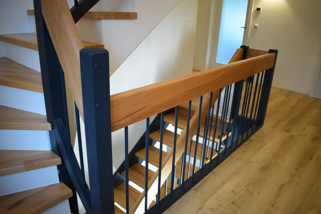 Moderne Innentreppe aus Holz mit schwarzem Metallgeländer, hellen Holzstufen und einem passenden Handlauf aus hellem Holz vor weißen Wänden und einem hellen Holzboden. Das Design ist sauber und zeitgemäß.