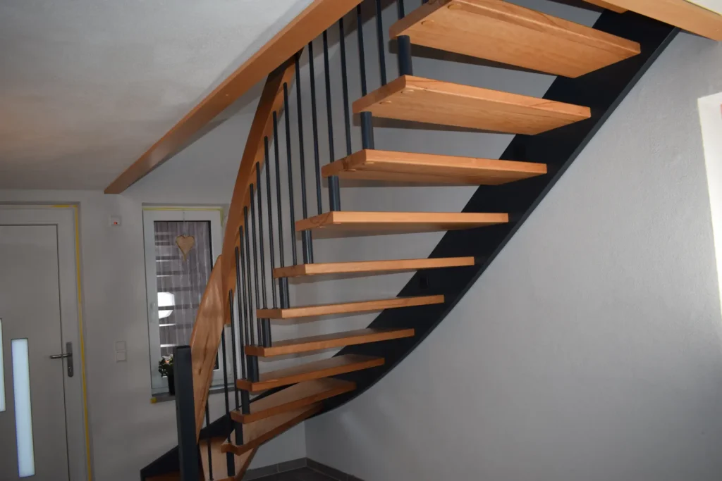Eine moderne Innentreppe mit Holzstufen und schwarzem Metallgeländer, die an einer weißen Wand befestigt ist. Das Design ist offen und minimalistisch und lässt das Licht durch die Lücken zwischen den Stufen hindurch.