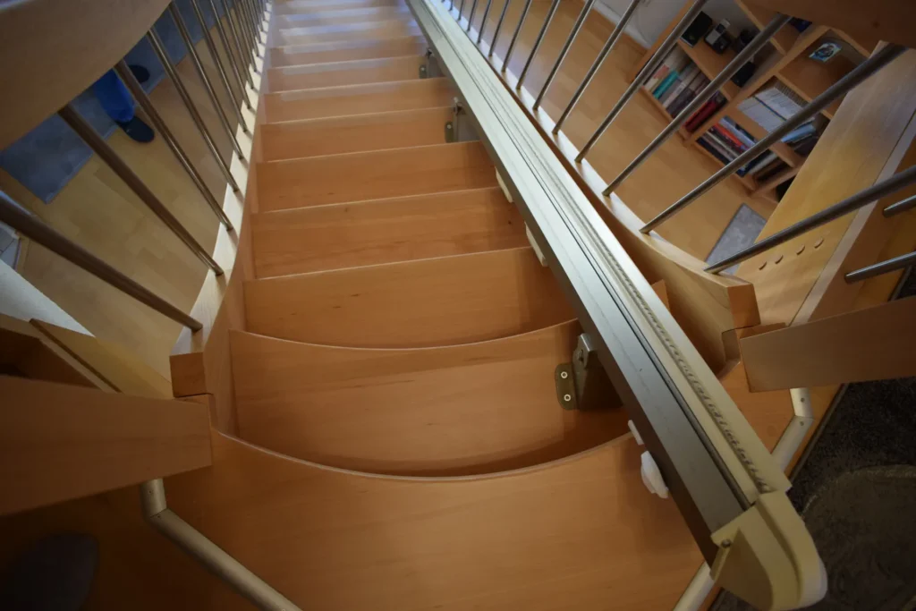Eine Holztreppe mit einem Metallhandlauf und einer an einer Seite angebrachten Treppenliftschiene, von oben gesehen mit Blick in einen Raum mit Regalen und Büchern.