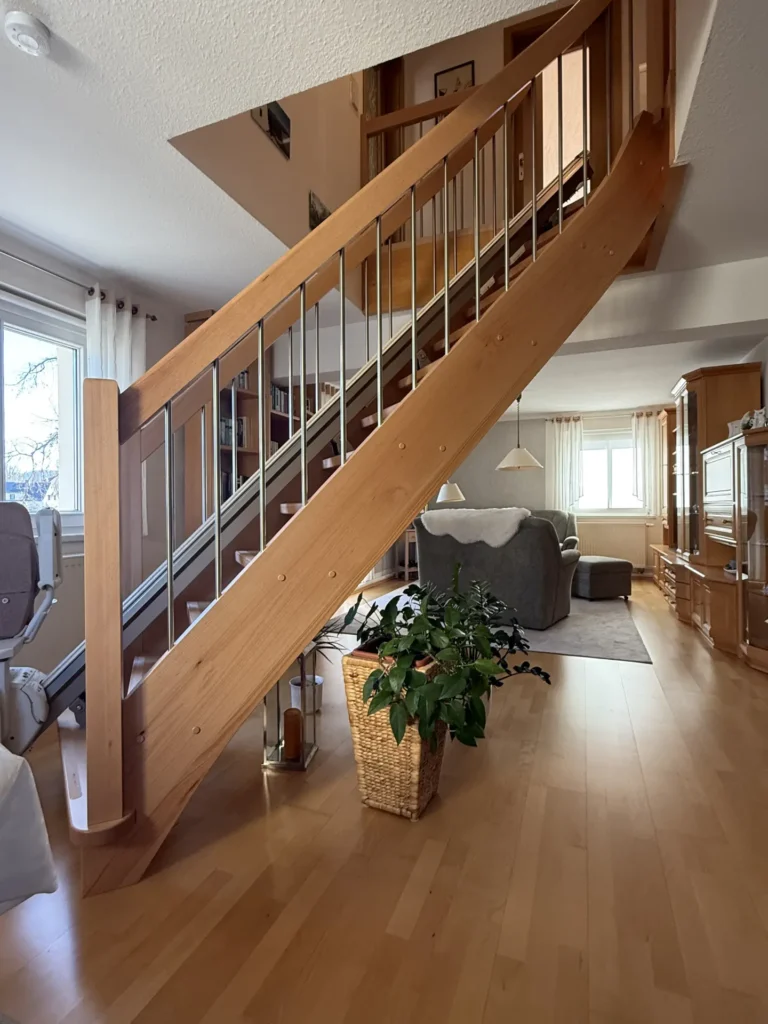Eine Holztreppe mit Metallgeländer führt ins Obergeschoss in ein helles Wohnzimmer mit Holzboden, einer grünen Topfpflanze unter der Treppe und großen Fenstern, die viel Tageslicht hereinlassen.