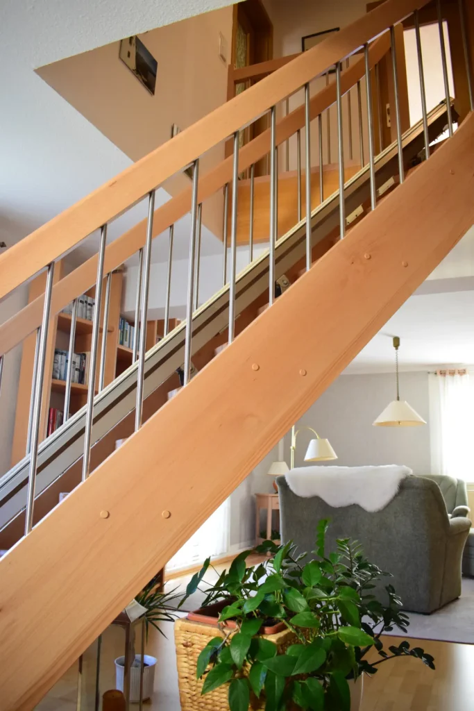 Eine Holztreppe mit Metallgeländer führt die Treppe hinauf in ein gemütliches Wohnzimmer. Eine grüne Topfpflanze steht in einem Korb unter der Treppe. Im Hintergrund stehen Bücherregale, eine Couch und Hängelampen an einem Fenster.
