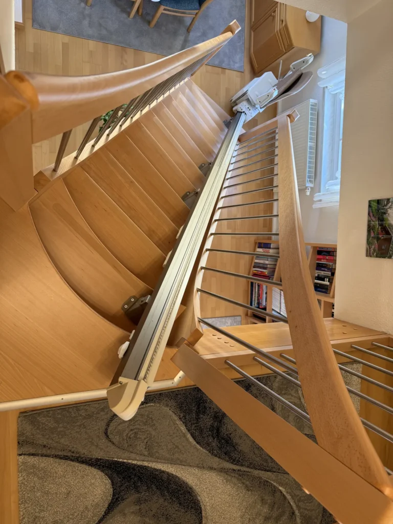 Eine Holztreppe mit einem Treppenlift, der am Seitengeländer angebracht ist. Die Treppe ist am unteren Ende gebogen und der Bereich ist durch ein Fenster in der Nähe gut beleuchtet. Am Fuße der Treppe befinden sich ein gemusterter Teppich und Bücherregale.