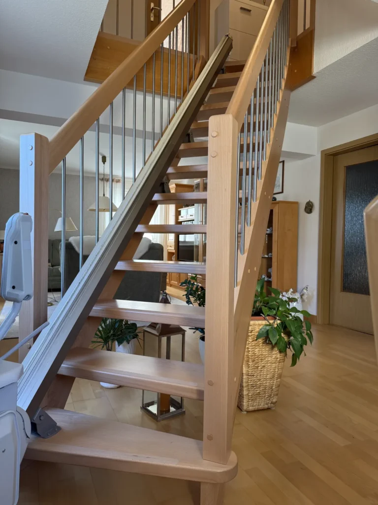 Eine Holztreppe mit Metallgeländer ist mit einem Treppenlift ausgestattet, der an den Stufen entlangführt. Die Umgebung ist mit Hartholzböden, Zimmerpflanzen und Holzmöbeln in einem hellen Wohnbereich ausgestattet.