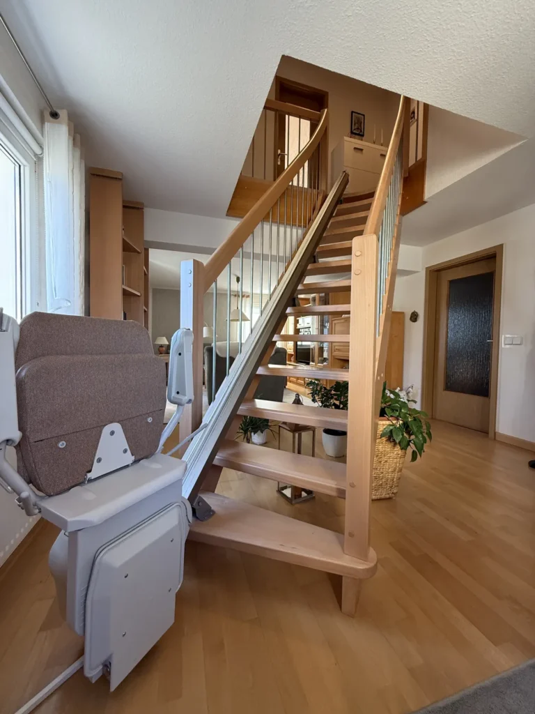 Eine hölzerne Wendeltreppe mit einem seitlich angebrachten Treppenlift, die in den zweiten Stock eines modernen, hellen Hauses mit Pflanzen und Holzmöbeln führt.