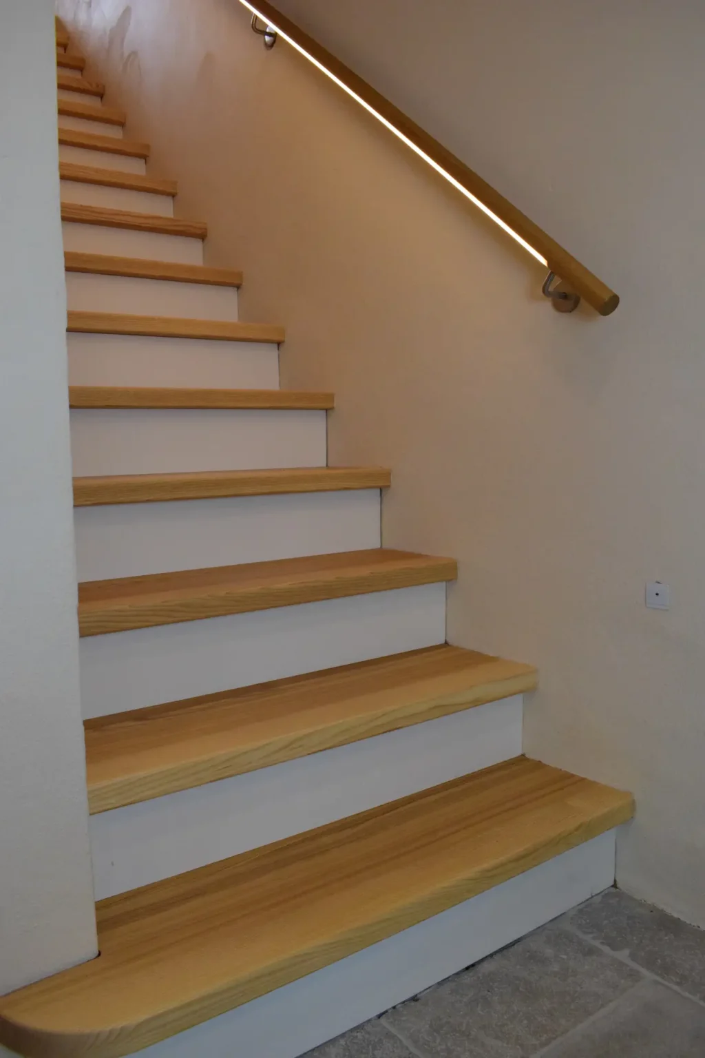 Holztreppe mit hellen Stufen und weißen Setzstufen, die entlang einer beigen Wand nach oben führt, mit einem Handlauf aus Holz und einem LED-Lichtband unterhalb des Handlaufs zur Beleuchtung.