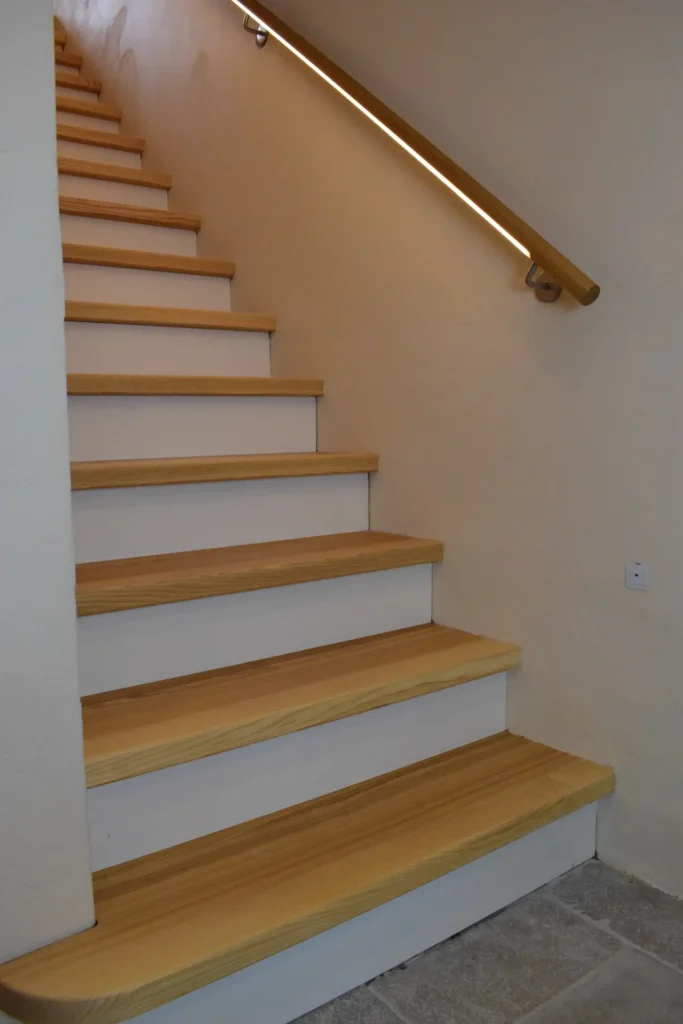 Holztreppe mit hellen Stufen und weißen Setzstufen, die entlang einer beigen Wand nach oben führt, mit einem Handlauf aus Holz und einem LED-Lichtband unterhalb des Handlaufs zur Beleuchtung.