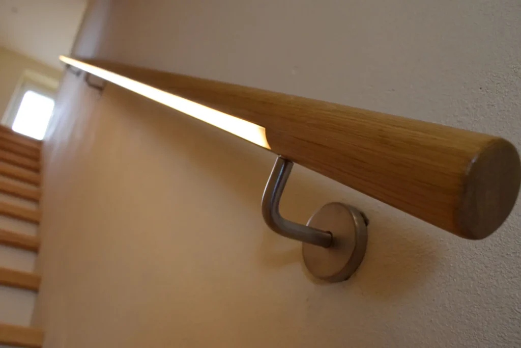 Nahaufnahme eines modernen Holzhandlaufs mit integrierter Lichtleiste, der an einer Wand neben einer Treppe angebracht ist und zu einem hellen Fenster im Hintergrund hinaufführt.