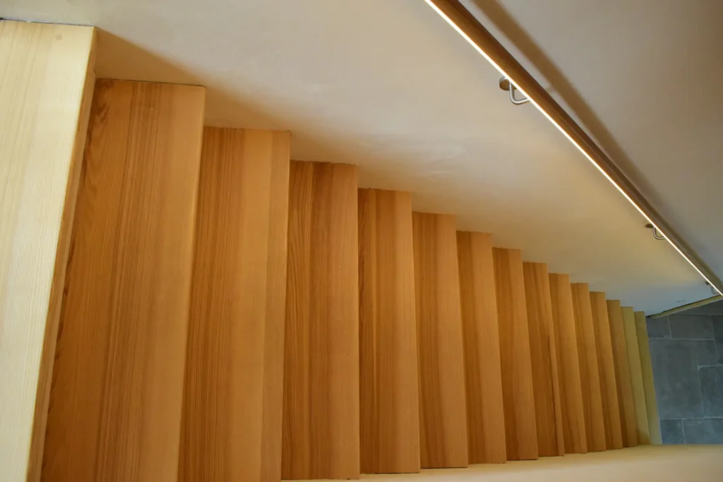 Eine Nahaufnahme einer modernen Holztreppe von oben, die das vertikale Muster der Stufen und eine parallel zur Treppe verlaufende lineare Deckenleuchte zeigt.
