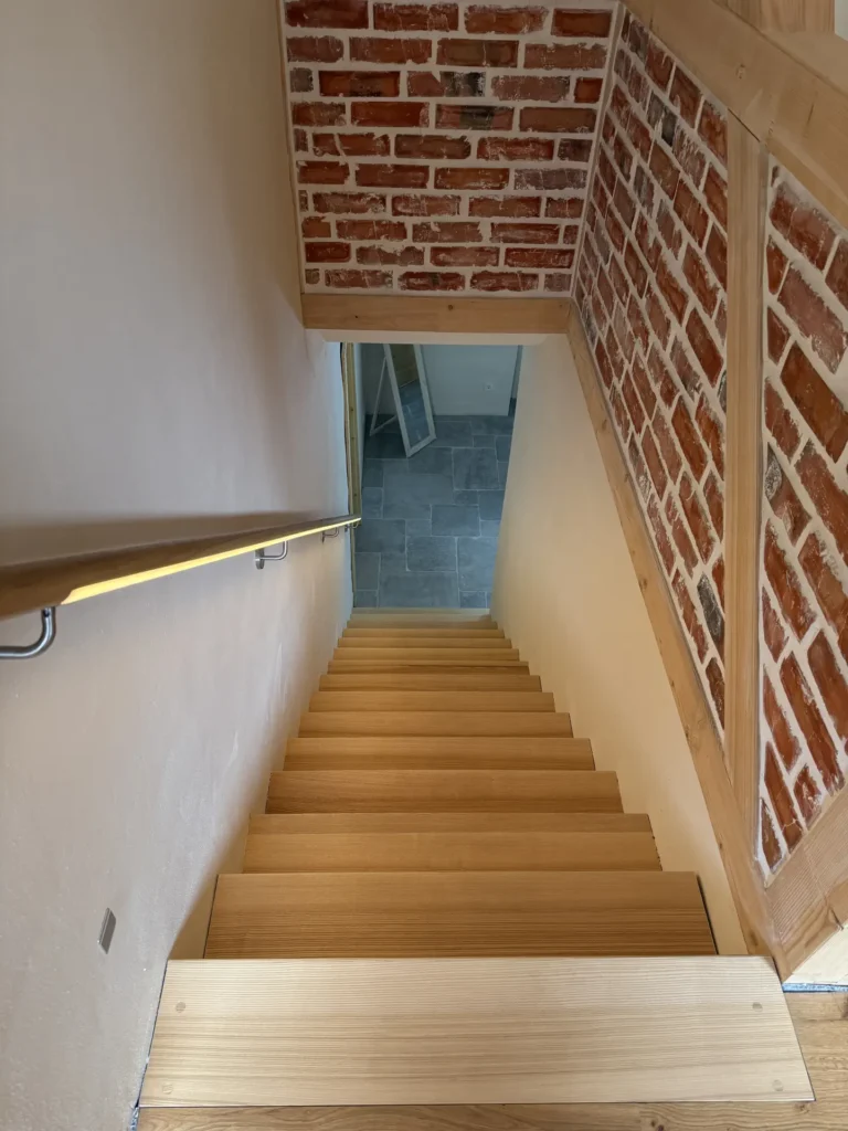 Eine Holztreppe mit Handlauf auf der linken Seite führt hinunter zu einem grau gefliesten Boden. Die rechte Wand und die Decke sind mit rotem Backstein verkleidet, was einen rustikalen Eindruck vermittelt. Der Raum ist durch natürliches Licht gut beleuchtet.
