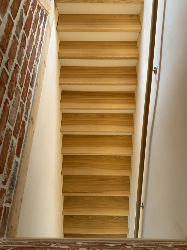 Holztreppe von unten gesehen, eingerahmt von weißen Wänden auf der rechten Seite und einer roten Ziegelwand auf der linken Seite. Auf dem Foto ist die Unterseite der Treppe zu sehen, die die Struktur und Maserung der Holzstufen zeigt.