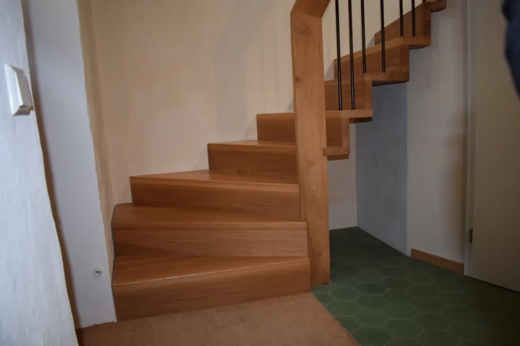 Eine Holztreppe mit eckigen Stufen biegt an einer Ecke nach rechts ab. Die Treppe befindet sich neben einer Wand mit einem Lichtschalter, und der Boden geht von Teppich in grüne sechseckige Fliesen über. Auf der rechten Seite ist ein schwarzes Geländer zu sehen.