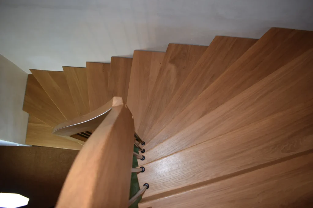 Blick von oben auf eine hölzerne Wendeltreppe mit geschwungenem Handlauf und dunklen Metallgeländern, die vor einer weißen Wand nach unten führt.