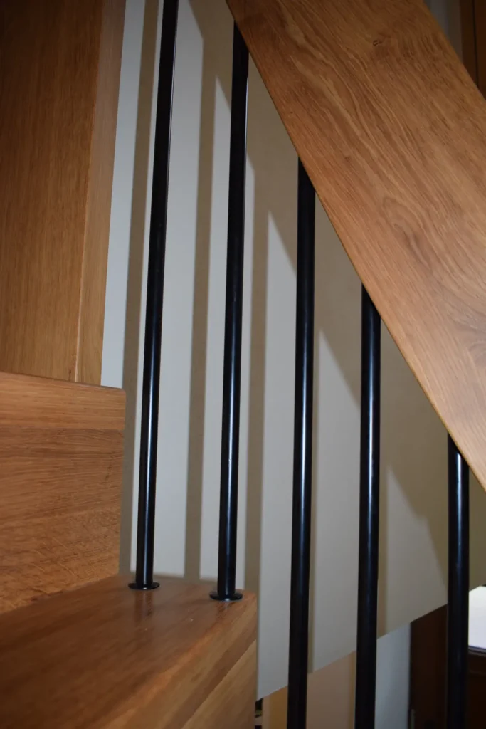 Nahaufnahme einer Holztreppe mit vertikalen schwarzen Metallgeländerstäben und hellen Wänden im Hintergrund. Das Foto konzentriert sich auf die Details der Holzmaserung und das Geländerdesign.