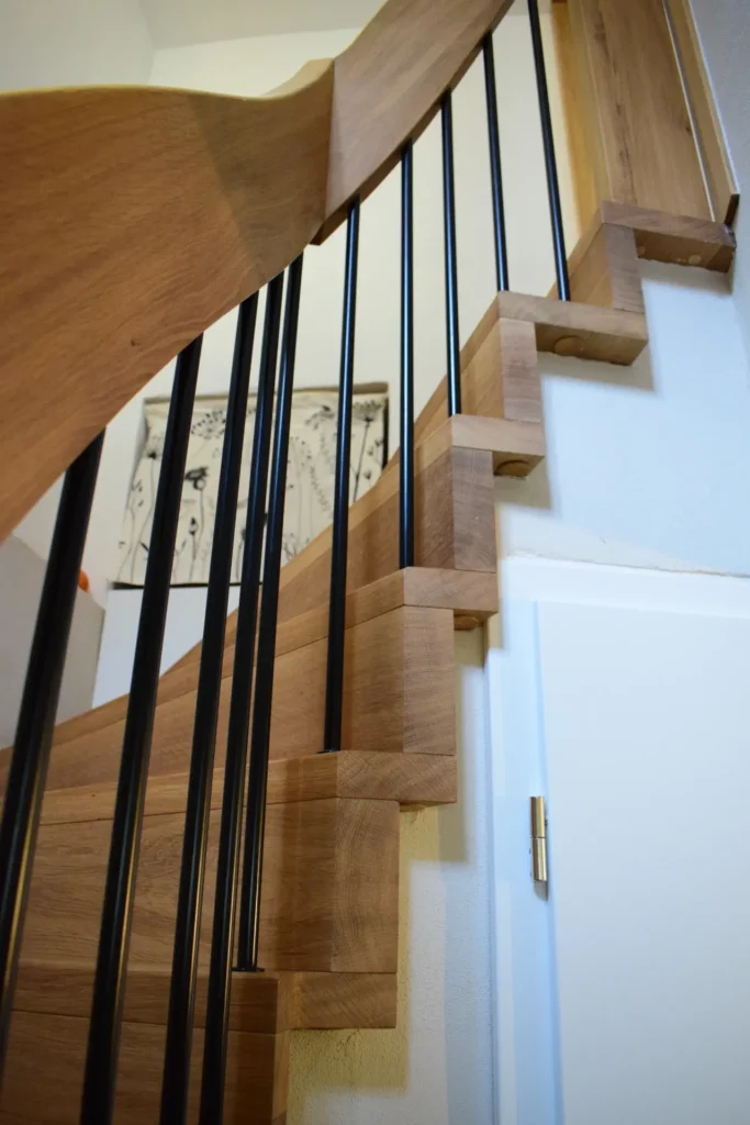 Eine Holztreppe mit offenen Setzstufen und schwarzen Metallgeländerstäben, die sich neben einer weißen Wand und Tür nach oben wölbt. An der Wand im Hintergrund ist ein gerahmtes Kunstwerk zu sehen.