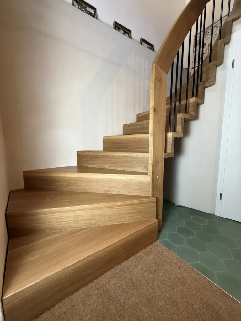 Eine moderne Holztreppe mit gewinkelten Stufen und einem geschwungenen Geländer, daneben eine Wand und ein Boden mit grünen sechseckigen Fliesen. Schwarze vertikale Baluster stützen das Geländer darüber.