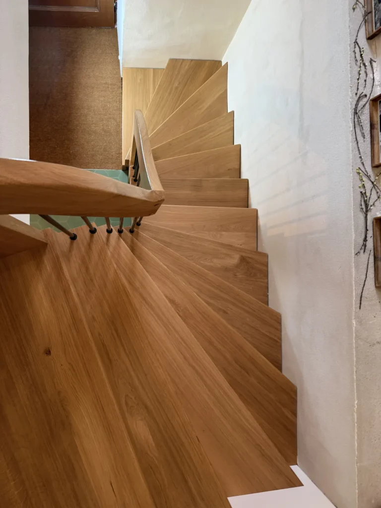 Draufsicht auf eine Holztreppe mit geschwungenem Handlauf, die zu einem Treppenabsatz hinunterführt. Die Treppe hat einen hellbraunen Anstrich und wird auf einer Seite von einer weißen Wand begrenzt.