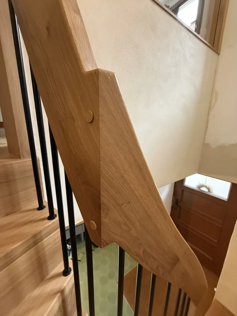 Eine Nahaufnahme einer modernen Holztreppe mit einem glatten Handlauf aus hellem Holz, vertikalen schwarzen Geländerstäben und einer Holztür am unteren Ende der Treppe. Die Wände sind cremefarben gestrichen.