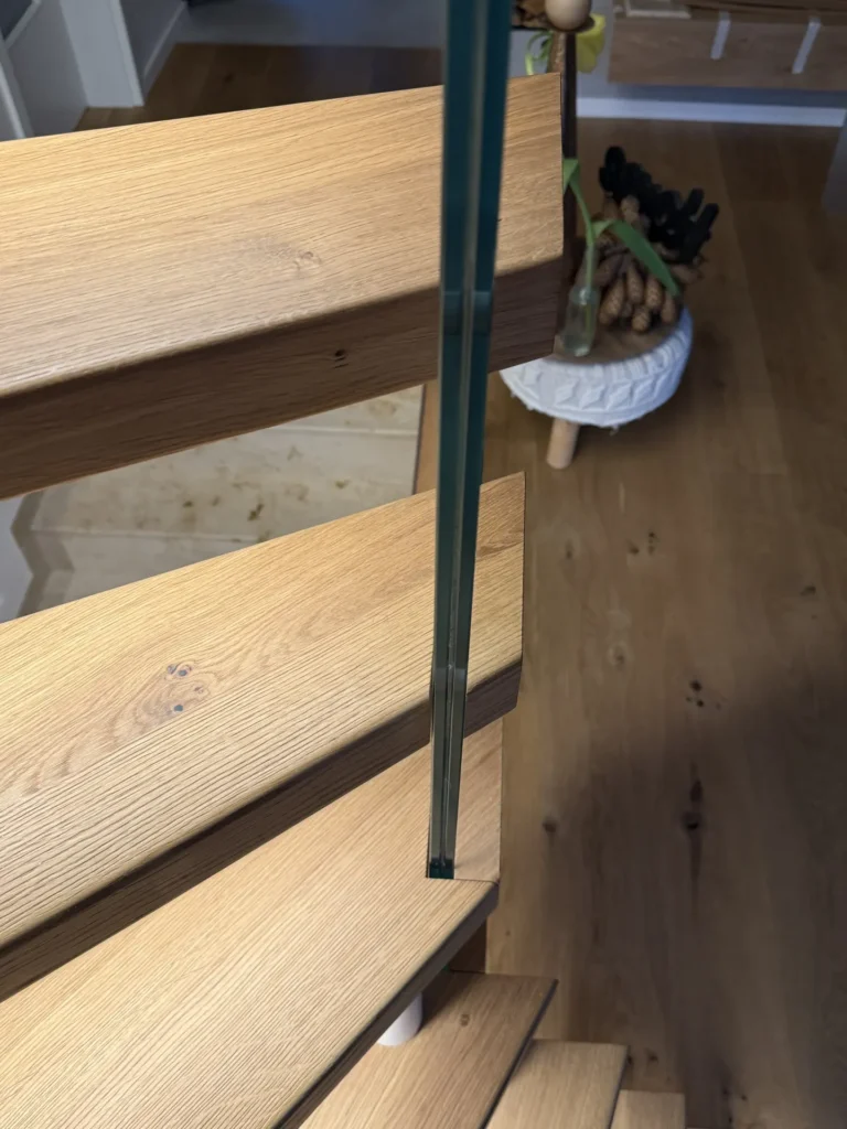 Nahaufnahme einer Holztreppe mit dünnem Glasgeländer von oben, Blick nach unten auf einen Holzboden und einen weißen Tisch mit einer Pflanze und einem Korb im Hintergrund.