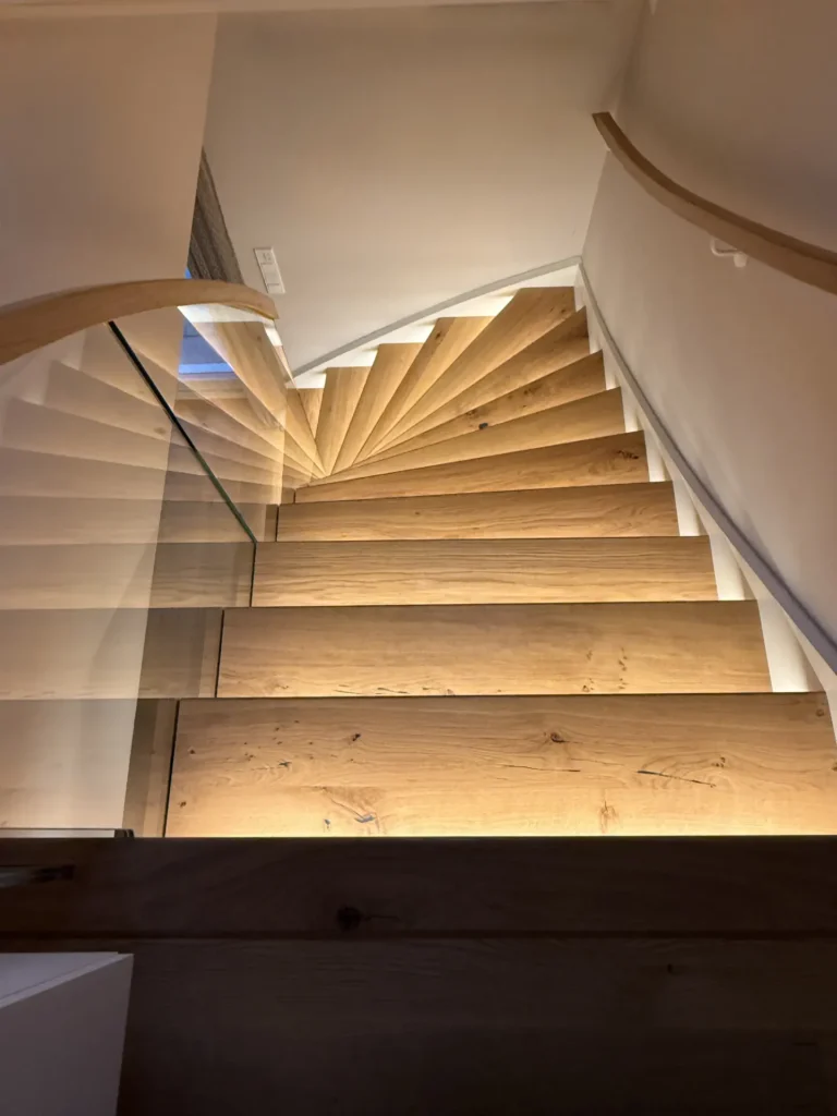 Eine Holzspindeltreppe mit warmer Beleuchtung unter den Stufen, geschwungenen Handläufen und einer Glasbalustrade, von oben betrachtet, in einer modernen Inneneinrichtung.