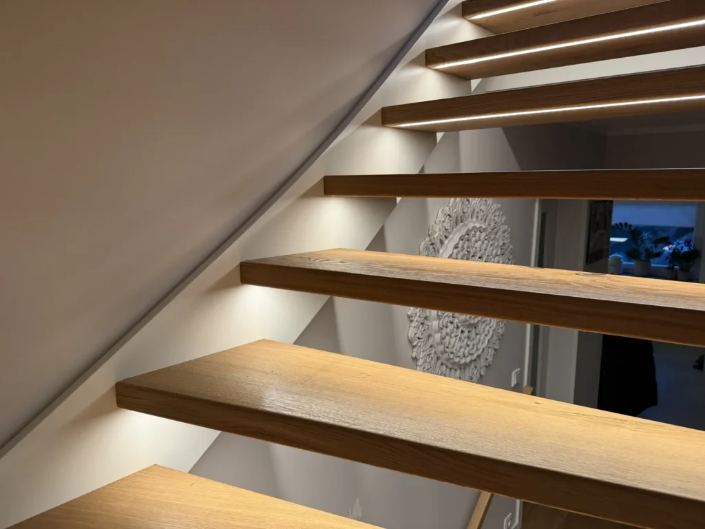 Schwebende Holztreppe mit eingebauter LED-Beleuchtung unter jeder Stufe, die an einer Wand befestigt ist. Unten ist ein moderner Flur mit einem dekorativen weißen Wandelement und einigen Zimmerpflanzen zu sehen.
