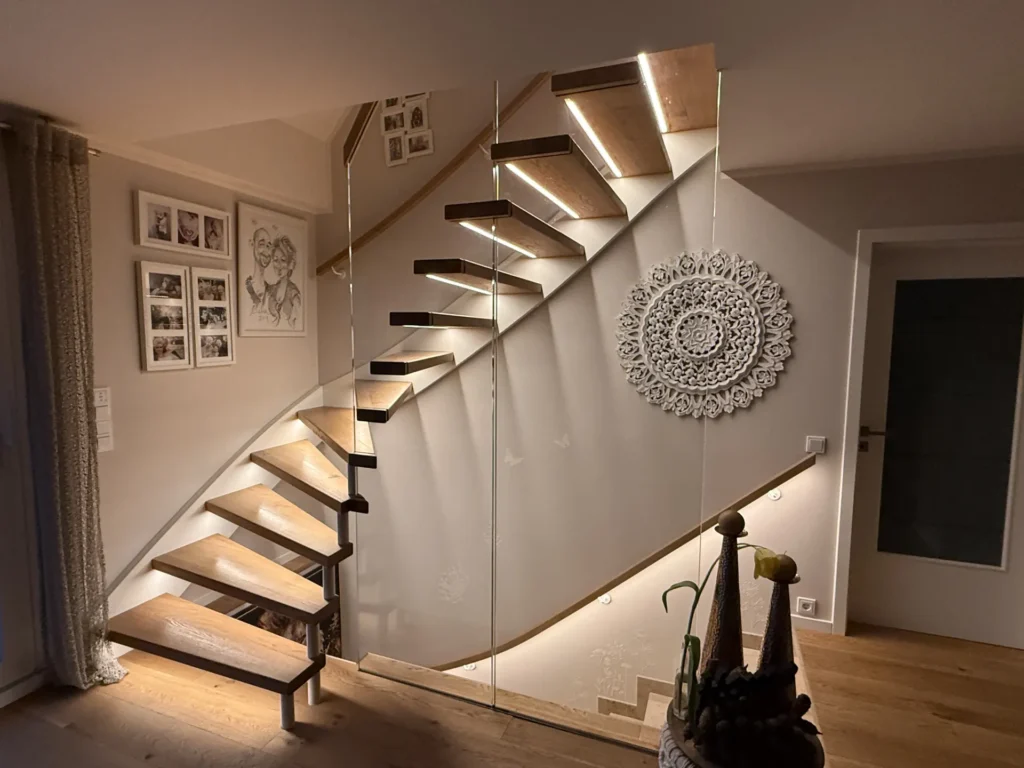 Eine moderne Holztreppe ohne Setzstufen, mit Glasgeländer und eingebauter Beleuchtung führt eine beigefarbene Wand hinauf, die mit gerahmten Fotos und einem großen, filigranen weißen Wandkunstwerk geschmückt ist. In der Nähe stehen Zimmerpflanzen auf dem Boden.
