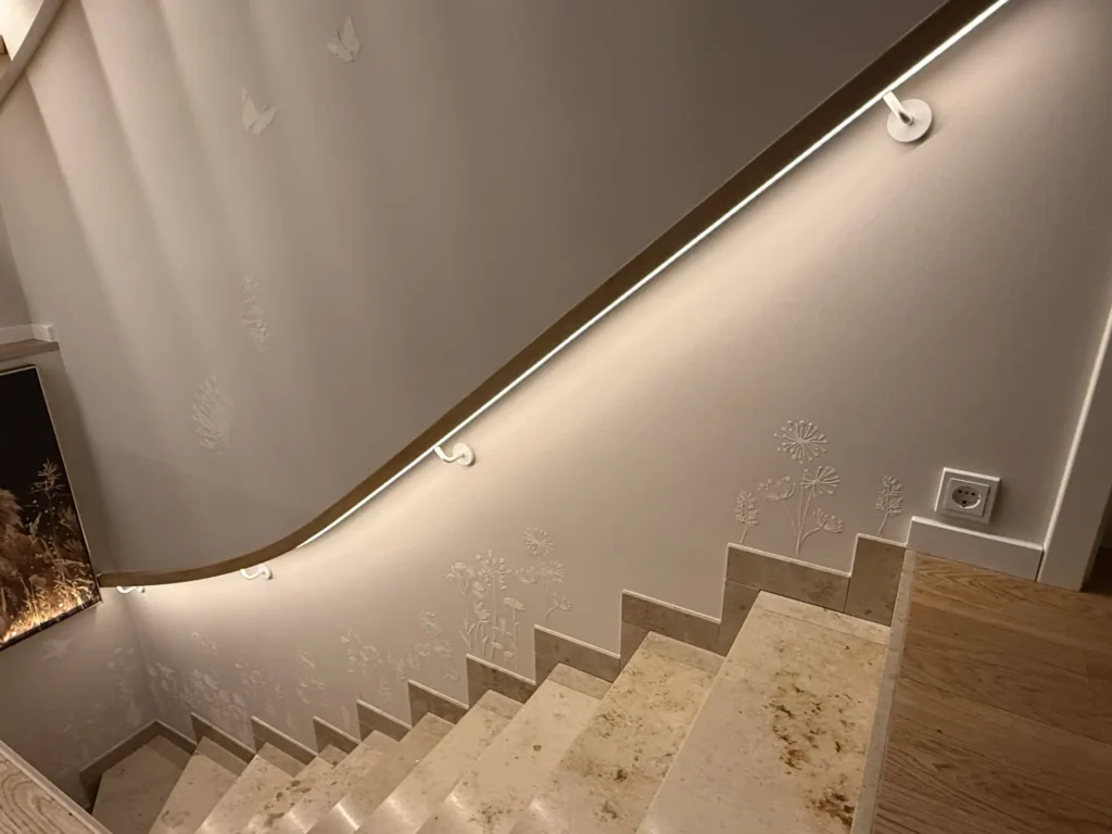 Eine gut beleuchtete Treppe mit beigen Marmorstufen, einem hölzernen Handlauf und dezenten Blumenmustern an der weißen Wand, beleuchtet von einem LED-Band entlang des Handlaufs.