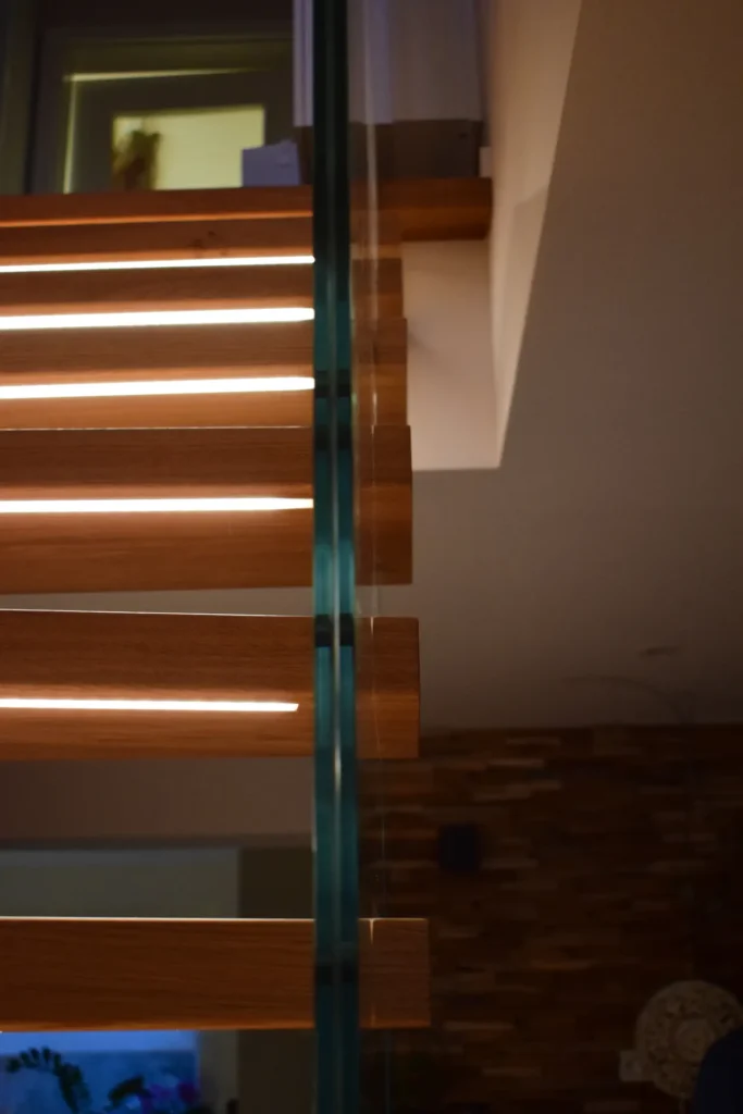 Eine moderne Holztreppe mit Glasgeländer, beleuchtet von eingebauten Leuchten auf jeder Stufe, in einem modernen Haus mit warmer Raumbeleuchtung.