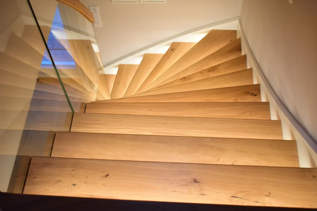 Eine moderne Holzspindeltreppe mit breiten, hellen Stufen und einem glatten Glasgeländer, von oben gesehen. Die Stufen werden von eingebauten Leuchten an der Wand beleuchtet, die eine warme und einladende Atmosphäre schaffen.