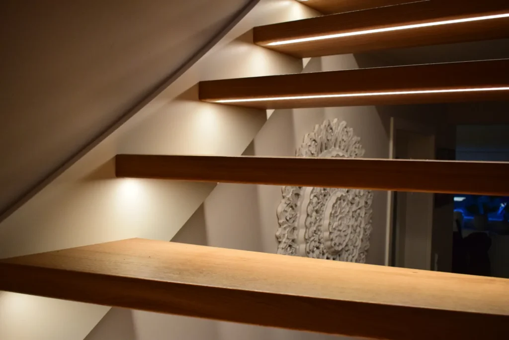 Nahaufnahme einer modernen schwebenden Holztreppe mit eingebauten LED-Lichtbändern an den Kanten, die ein warmes Licht auf die Wand werfen und ein dekoratives Reliefkunstwerk unter der Treppe hervorheben.