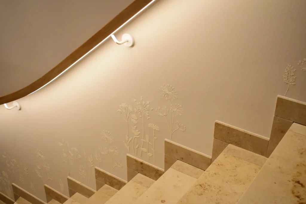 Nahaufnahme einer beigen Steintreppe mit Holzhandlauf und Wandleuchte. Die Wand ist mit geprägten floralen Mustern versehen, die dem minimalistischen Treppendesign ein dekoratives Detail hinzufügen.