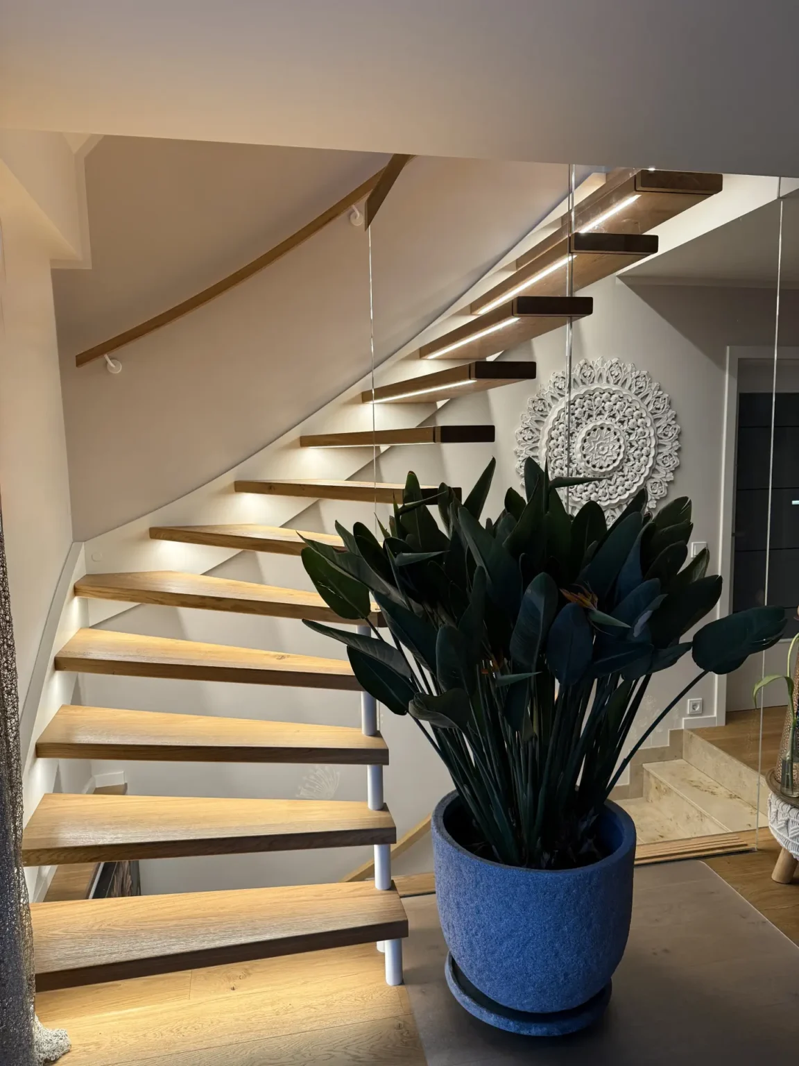 Eine moderne, geschwungene Holztreppe mit eingebauten Lampen führt neben einer großen Grünpflanze in einem blauen Topf nach oben. Dekorative weiße Wandkunst hängt an der neutralfarbenen Wand in einem stilvollen Interieur.