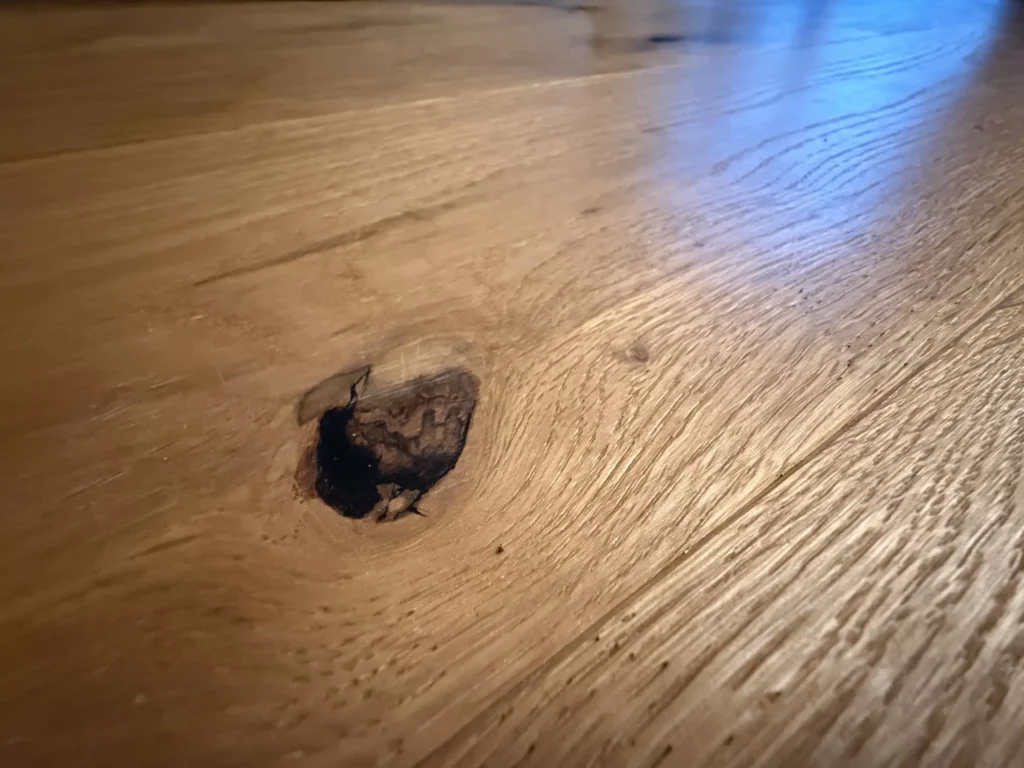 Nahaufnahme einer Holzoberfläche mit einem dunklen Knoten in der Maserung, umgeben von einer natürlichen Holzstruktur und einem warmen, braunen Farbton. Das Bild hebt das organische Muster und die Unebenheiten des Holzes hervor.