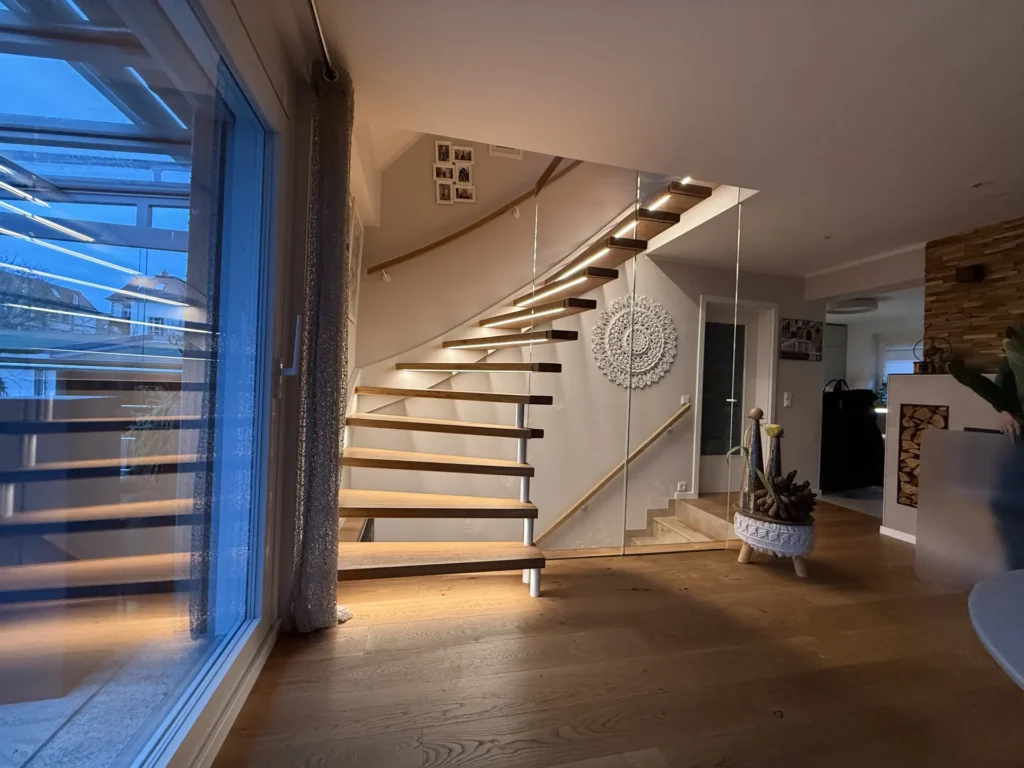 Modernes Interieur mit einer schwebenden Holztreppe mit Glasgeländer, einer dekorativen weißen Wand, Holzböden und einem großen Fenster, das blaues Abendlicht reflektiert. Eine Pflanze und kleine Fotos schmücken den Raum.