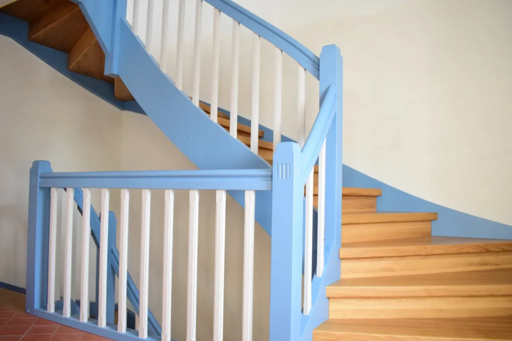 Eine Holztreppe mit hellbraunen Stufen und einem blauen Handlauf mit weißen vertikalen Balustern vor einer hellen Wand in einem Innenraum.