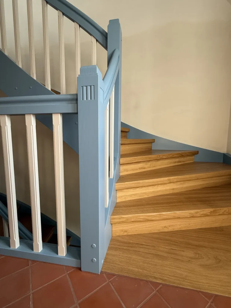 Eine Holztreppe mit hellbraunen Stufen und einem blau-weißen Geländer schlängelt sich neben einer cremefarbenen Wand nach oben, die über einem roten Fliesenboden liegt.